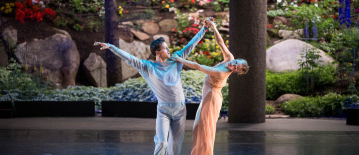 Robbie Fairchild and Sara Mearns perform on an open air stage with greenery in the background. Fairchild stands leaning into one hip, arms raised in a high V as he looks toward Mearns. Mearns holds his hand and shoulder while leaning away from him, balanced en pointe with one foot crossed over the other. They were simple, gauzy costumes, Fairchild in pale grey and Mearns in champagne.