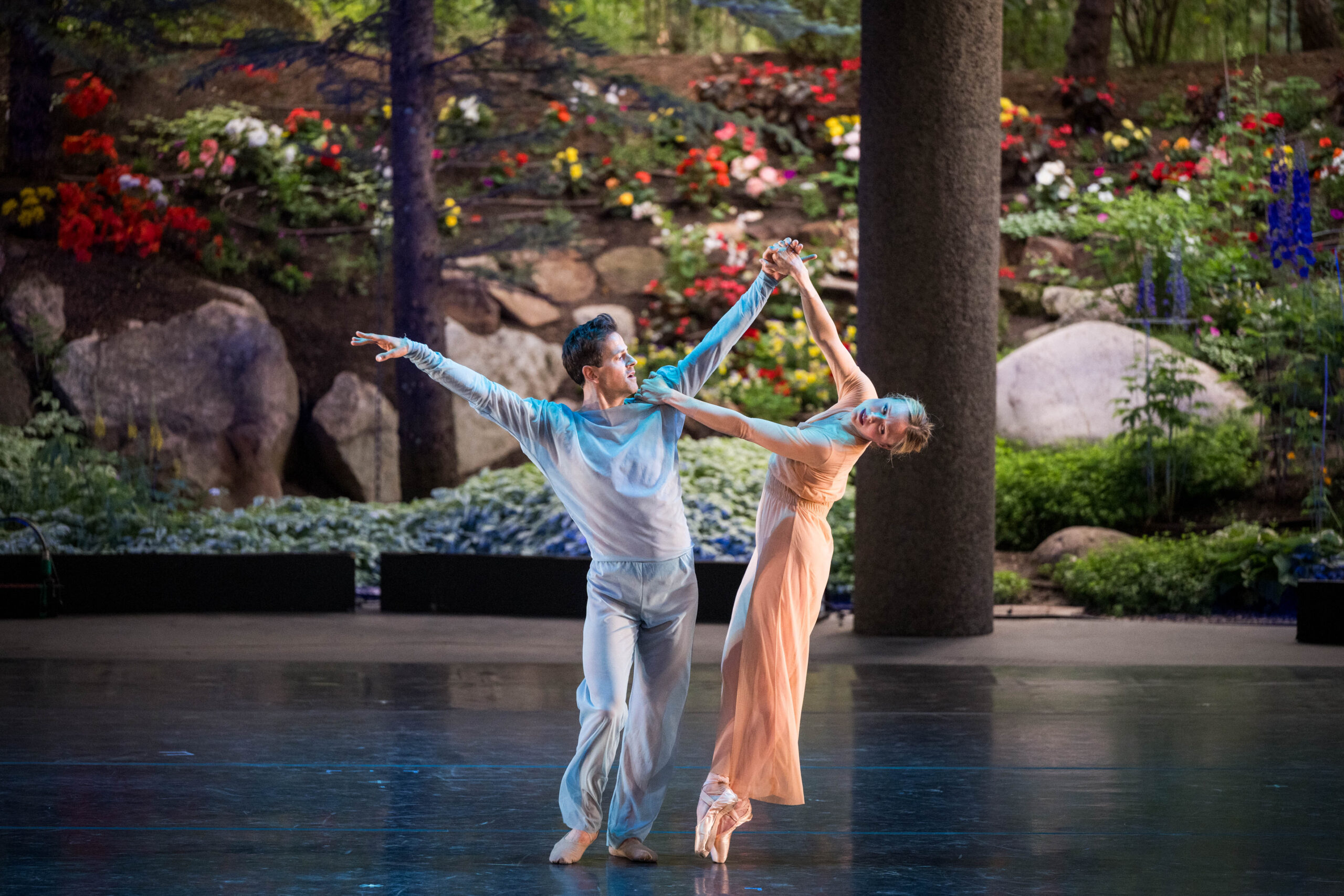 Robbie Fairchild and Sara Mearns perform on an open air stage with greenery in the background. Fairchild stands leaning into one hip, arms raised in a high V as he looks toward Mearns. Mearns holds his hand and shoulder while leaning away from him, balanced en pointe with one foot crossed over the other. They were simple, gauzy costumes, Fairchild in pale grey and Mearns in champagne.