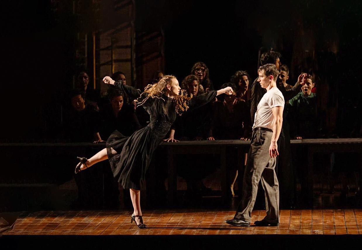 Payton Johnson performs an arabesque while reaching towards Alexander Bozinoff. She wears a long sleeve, knee-length black dress and character heels.