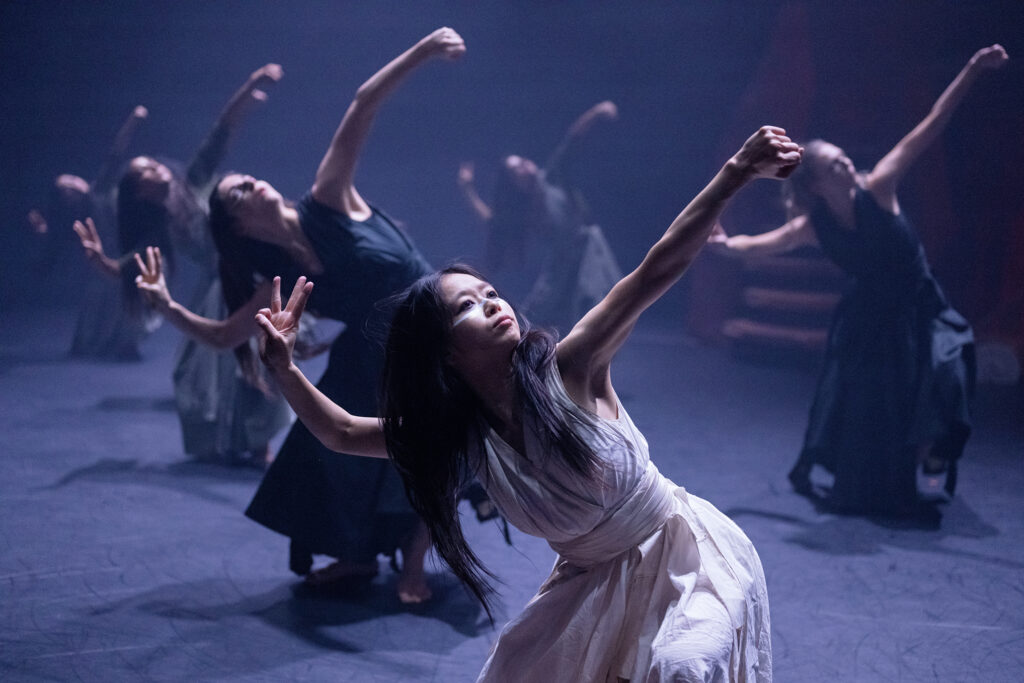In the foreground, a dancer bends forward and swings her left arm up on a diagonal, gaze following, while her left hand is held up beside her face with pinkie finger and thumb pressed together. She is costumed in white, with her hair loose around her shoulders. Five dancers wearing darker versions of the costume are hazy in the background as they move through the same gesture.