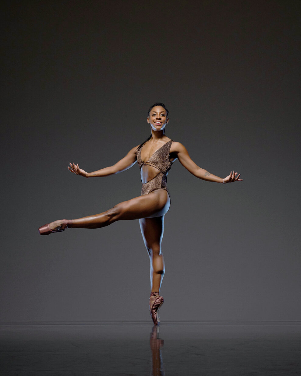 Isabela Coracy stands en ppinte with one leg extended devant and her palms up. She wears a skin tone leotard and brown pointe shoes.