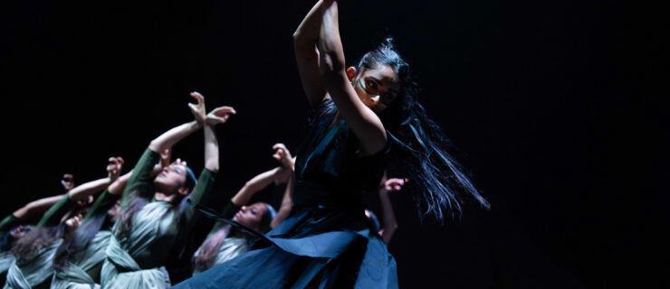 On a dark stage, a line of women in long dark blue and green dresses strike similar poses, with their hands over their heads, crossed delicately at the wrists.