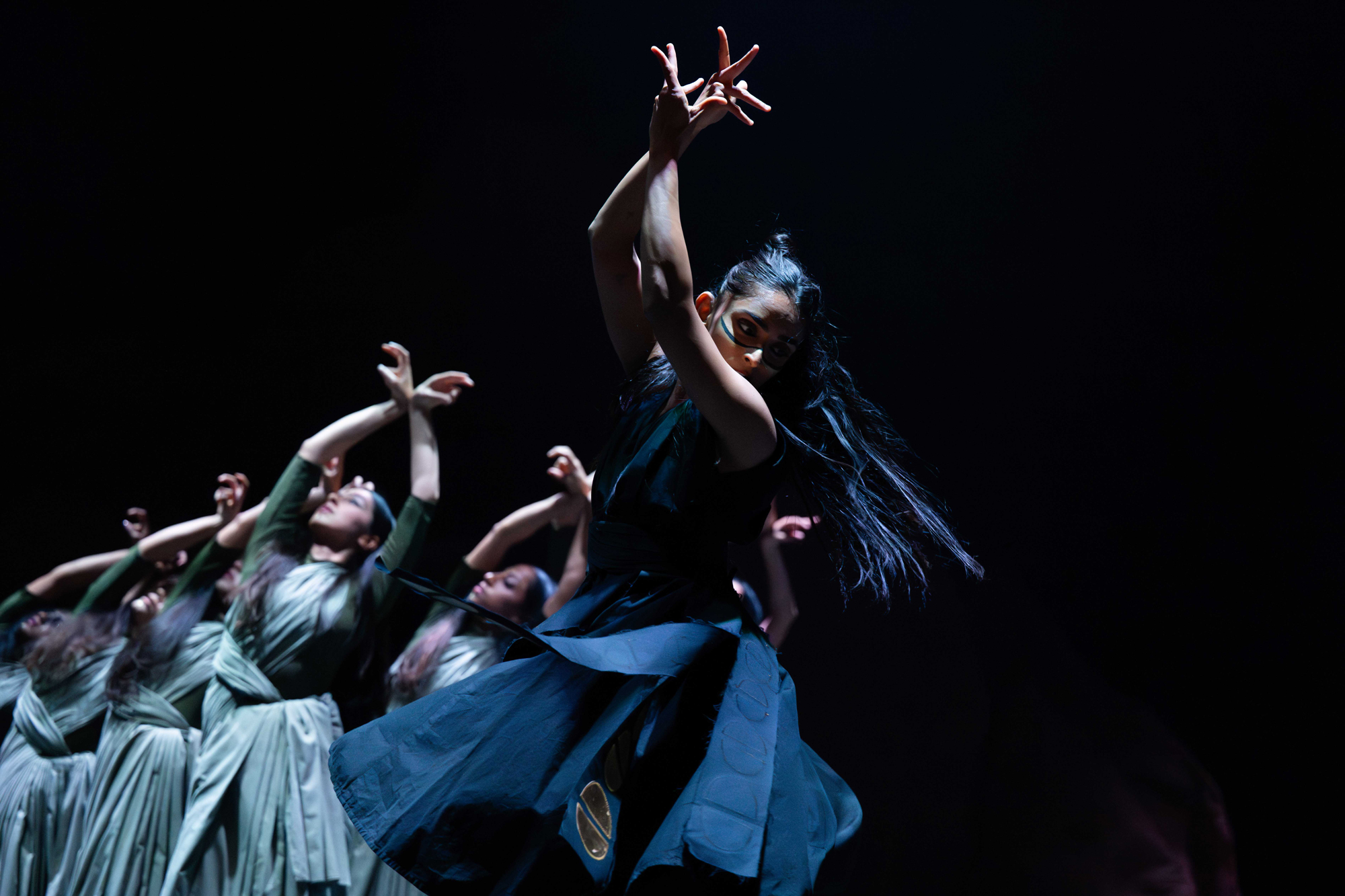 On a dark stage, a line of women in long dark blue and green dresses strike similar poses, with their hands over their heads, crossed delicately at the wrists.