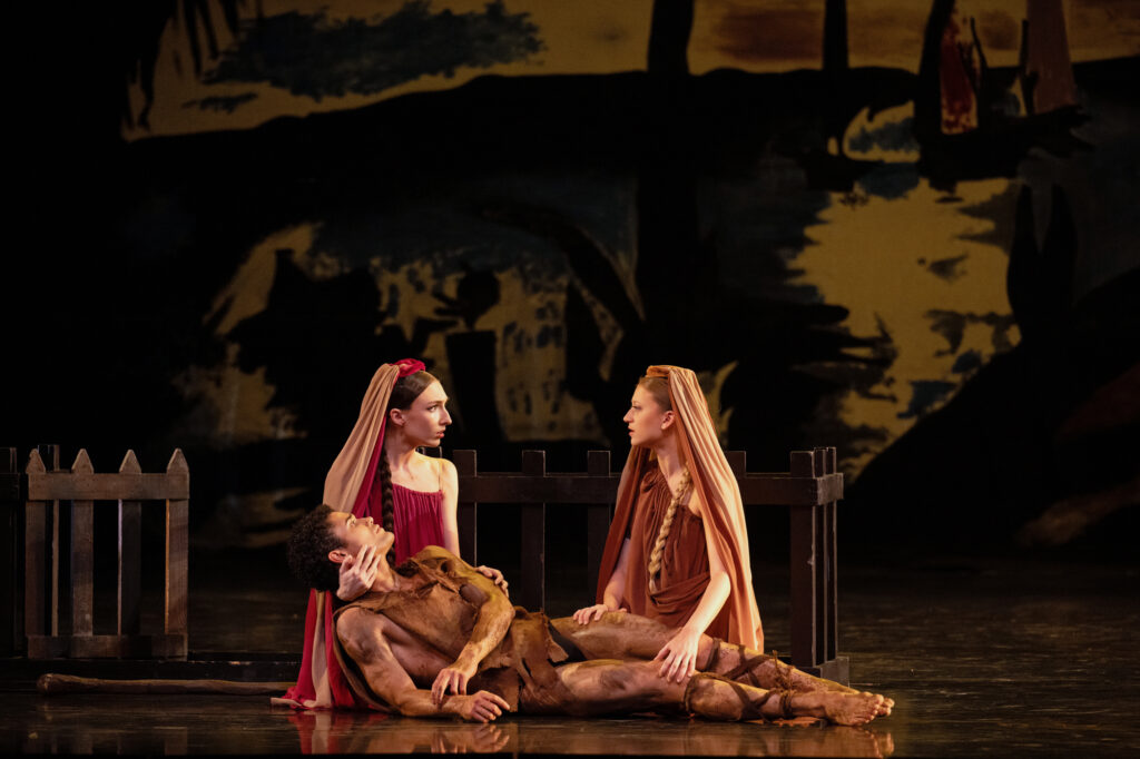 Sophie Poulain and Georgie Grace Butler hold Sayron Pereira as he lies on stage. They wear veils and red dresses.