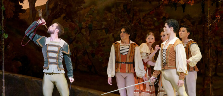 Carraig New blows a horn while Ángel Ramírez stands next to him with a sword. Corps dancers look on behind them. They wear medieval style costumes.