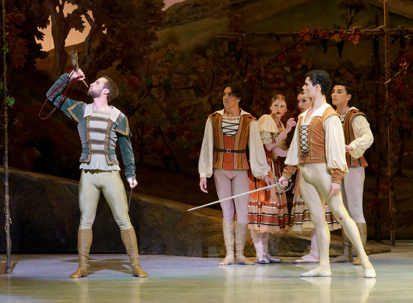 Carraig New blows a horn while Ángel Ramírez stands next to him with a sword. Corps dancers look on behind them. They wear medieval style costumes.