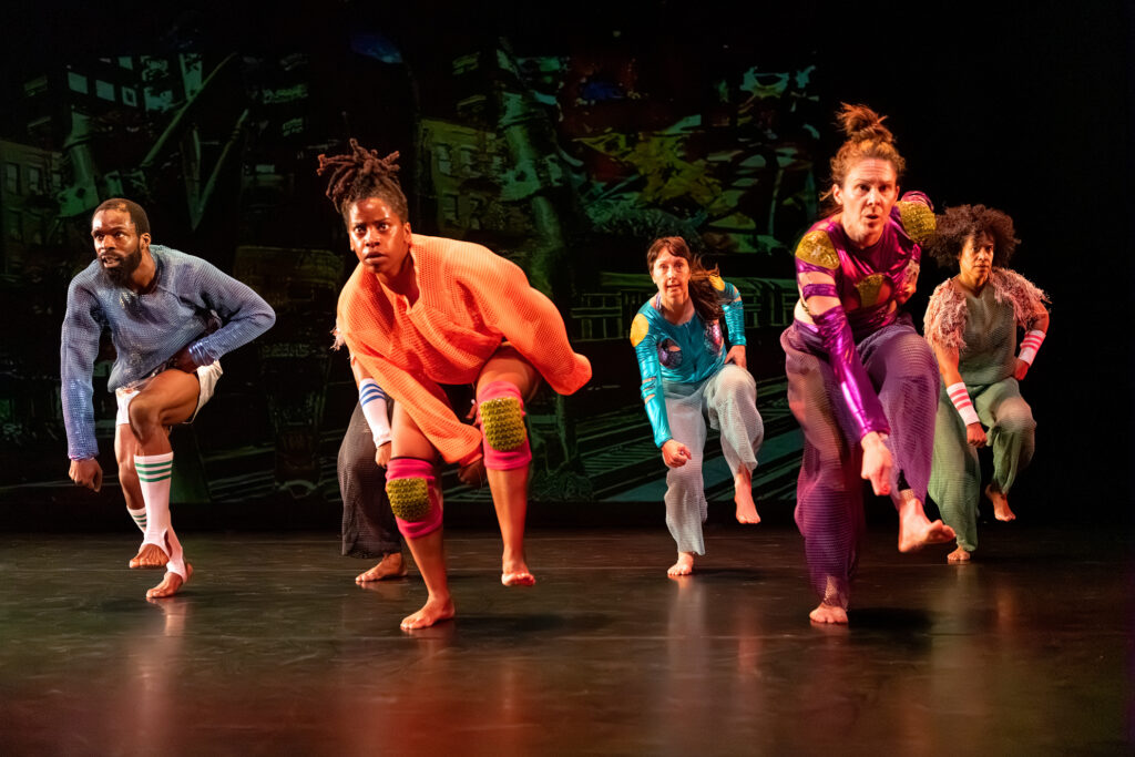 Five contemporary dancers perform a dynamic, grounded routine on a dark stage. They wear vibrant, eclectic outfits—including a textured orange sweater with green-studded knee pads, a metallic purple bodysuit, and a shimmering blue long-sleeve top. Their expressions are intense and focused as they balance on one leg or lean forward in athletic poses. The backdrop features a dark, abstract mural of urban and geometric shapes.