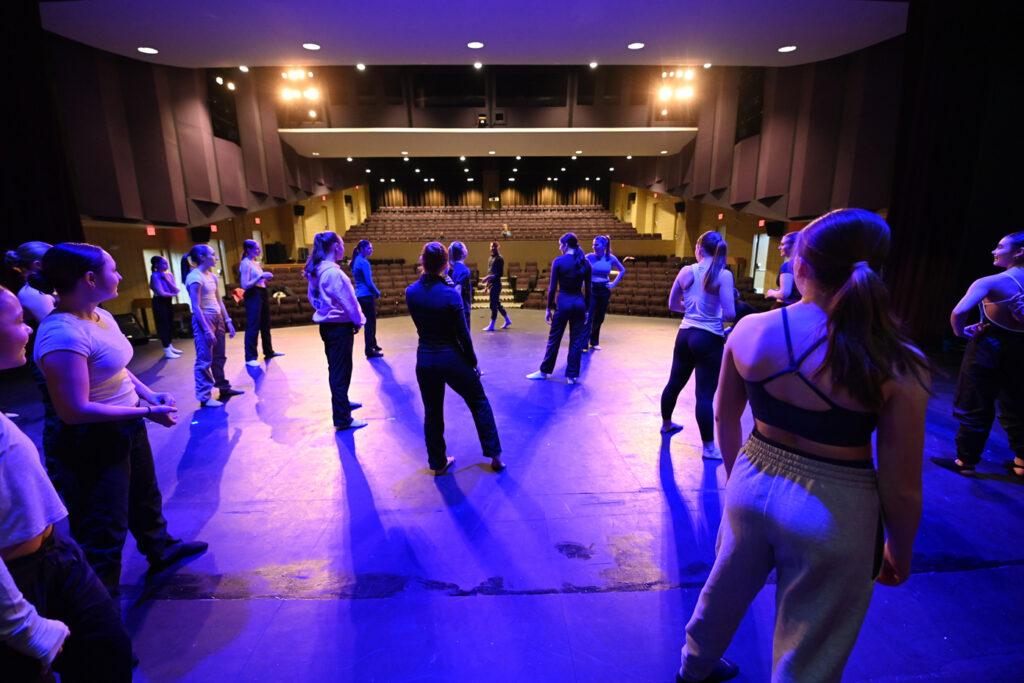 A large group of Saint Anselm College dancers stand onstage in a proscenium theater.