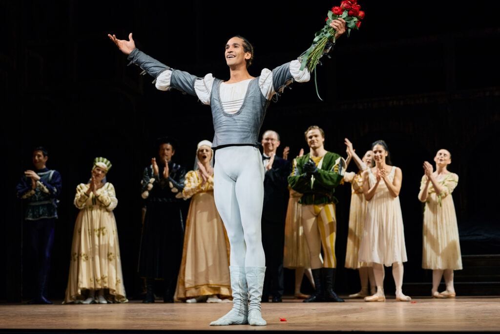 Davi Ramos takes a bow, holding a bouquet of roses aloft while his fellow company members applaud and cheer upstage.