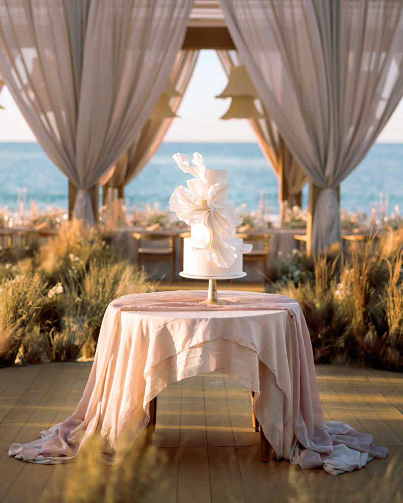 A white wedding cake with a large flowers sits on a table in front of the ocean.