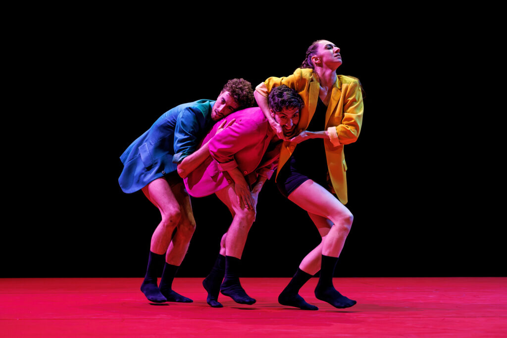 Three dancers wearing brightly colored suit jackets over black tank tops and shorts form a line. The one at the rear rests their head on the back of the dancer in front of them; the dancer in center smiles zanily as they stick their head through a window formed by the arms of the dancer in front.