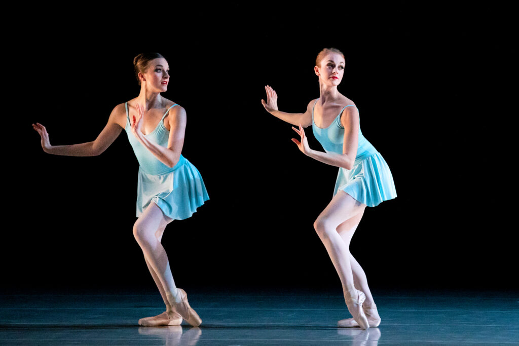 Jordan Fry and Kazlyn Nielsen wearing simple blue leotards and skirts. They pop one foot and lean forward with their palms pressing away.