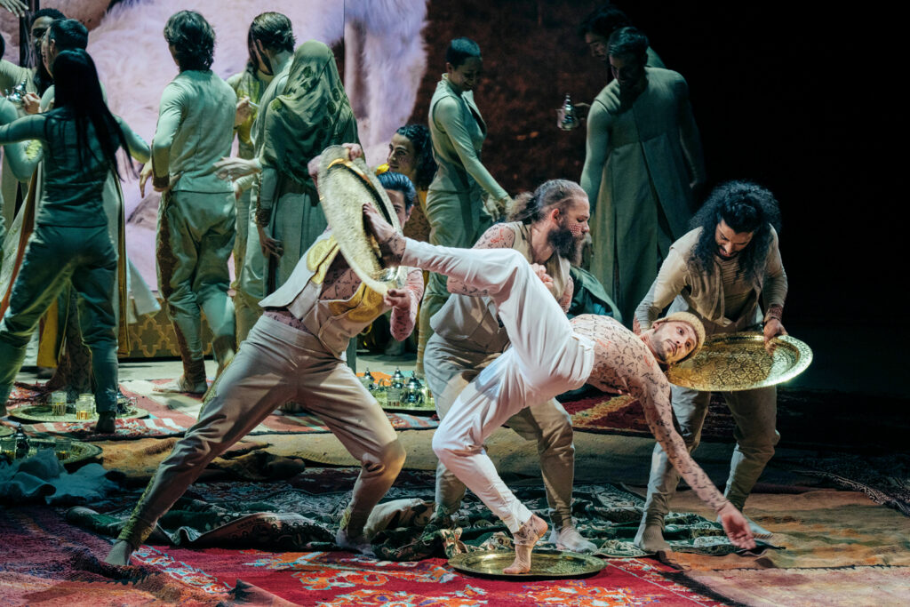 A dancer balances in a forced arch layout on a golden serving tray; another tray is held behind his head, a third to his upraised foot. A dancer supports him with one arm under his back. Rugs of different colors and patterns are laid out across the floor; upstage, numerous dancers cluster around tea services.