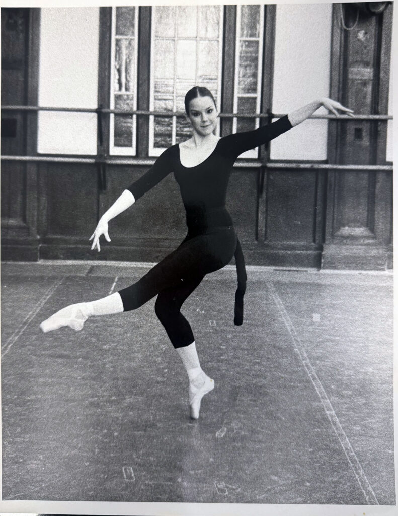 A black-and-white photo of a young Rice in a ballet studio. She wears a dark leotard, tights, and pointe shoes, standing on pointe on a hooked right foot as she extends her left leg in front of her.
