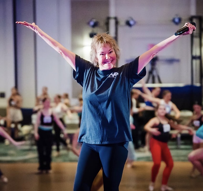 Rice, wearing a dark t-shirt and holding a microphone in her left hand, is pictured onstage in a dance convention ballroom, her arms extended in a high V, smiling broadly.