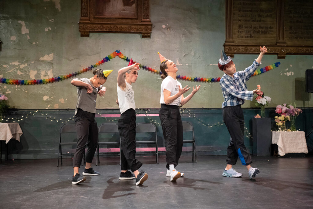 Four dancers in party hats dance like they're at a party in a line in front of a row of folding chairs.