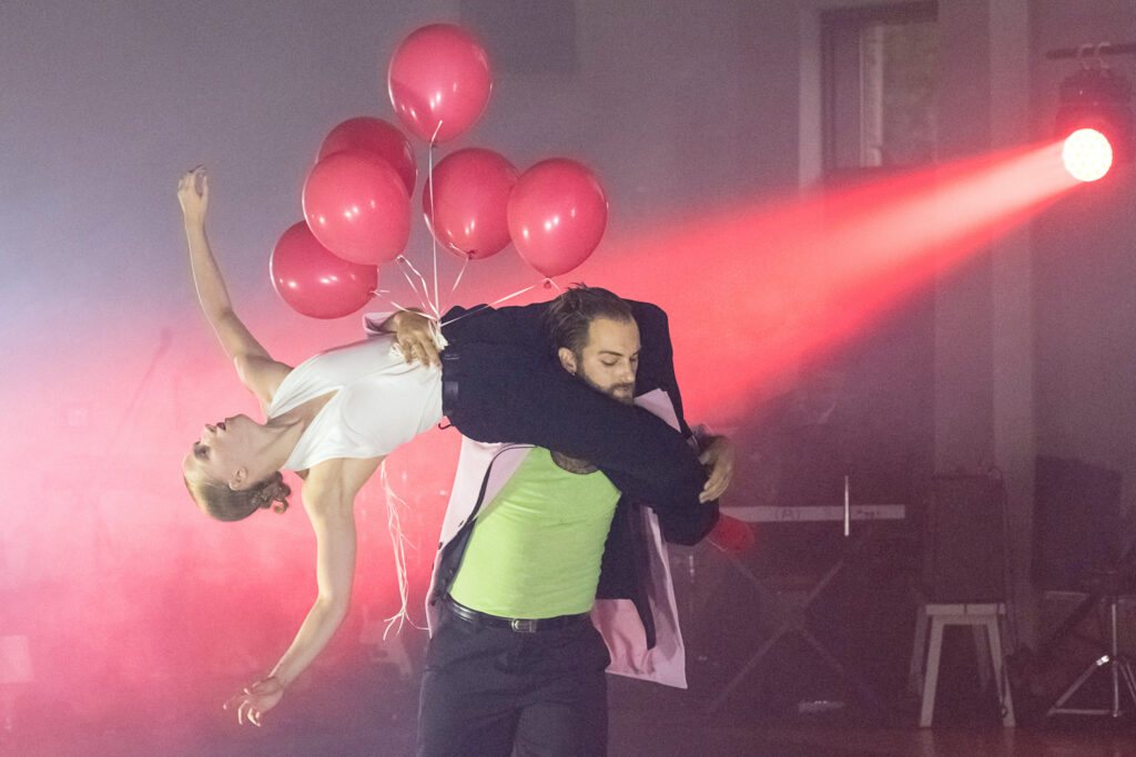 A dancer wearing a lime green tank carries another over their shoulders, the hand wrapped around the lifted dancer's torso holding a cluster of pink balloons. The lifted dancer arches back to look up to the sky.