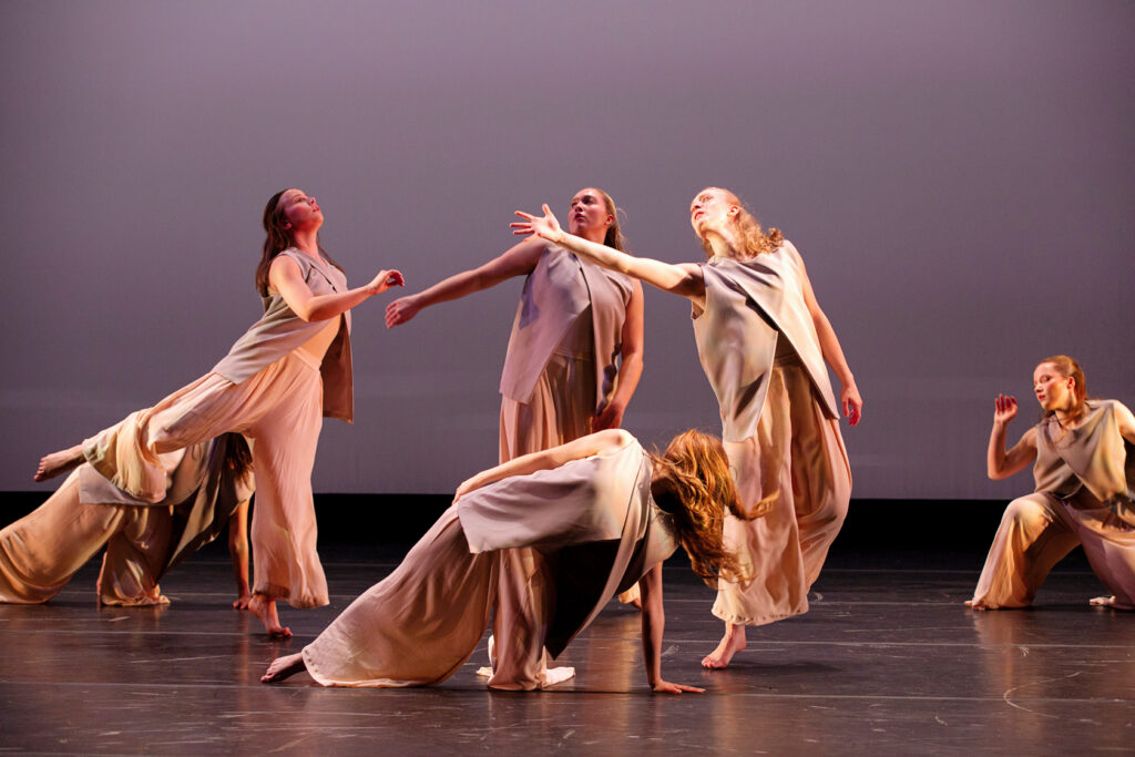 Samford University students performing on stage wearing tan tunics and wide-legged pants. Some crouch on the floor while others stand and reach.