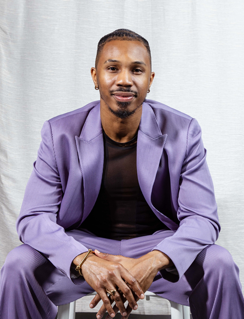 O'Meally, wearing a purple suit, sits on a stool in front of a white dropcloth, his hands crossed in front of him, a slight smile on his face.