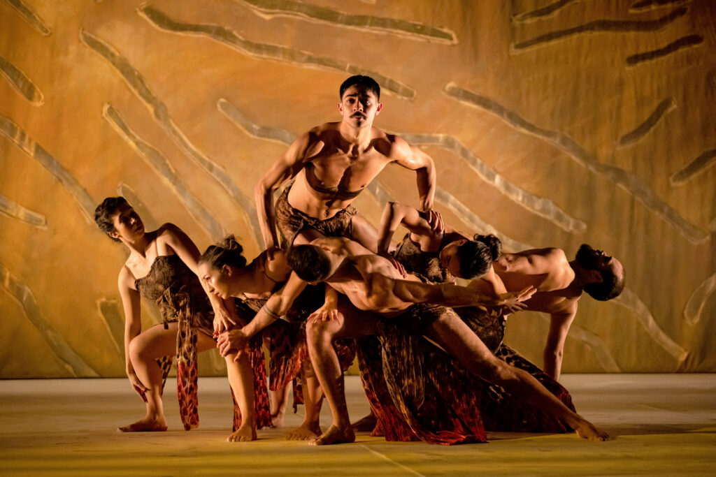 A bare-chested dancer gazes out over a low wall formed by five dancers who interlock and interweave as they lunge and crouch. The stage is awash in a dusty yellow.