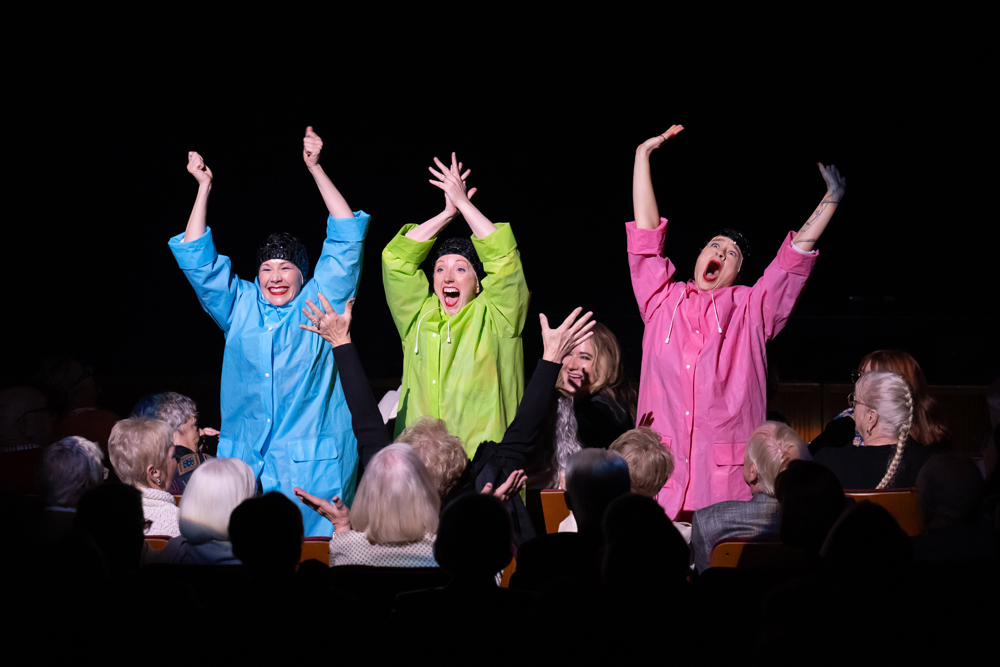 Wilson, Meyers, and Lawson stand between rows of seats in a darkened auditorium, their arms flung over their heads, their faces goofily joyous. They wear matching raincoats—Wilson's is blue, Meyers' is green, and Lawson's is pink.