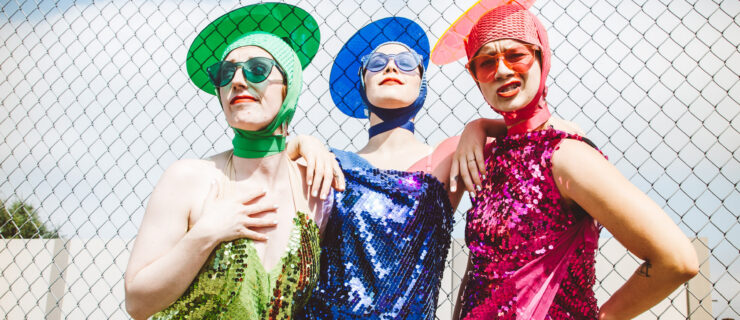 The three Seaweed Sisters strike quirky poses outdoors, backed by a blue sky. They wear sequined dresses, sunglasses, and disc-like hats; Meyers' ensemble is green, Wilson's is blue, and Lawson's is pink.