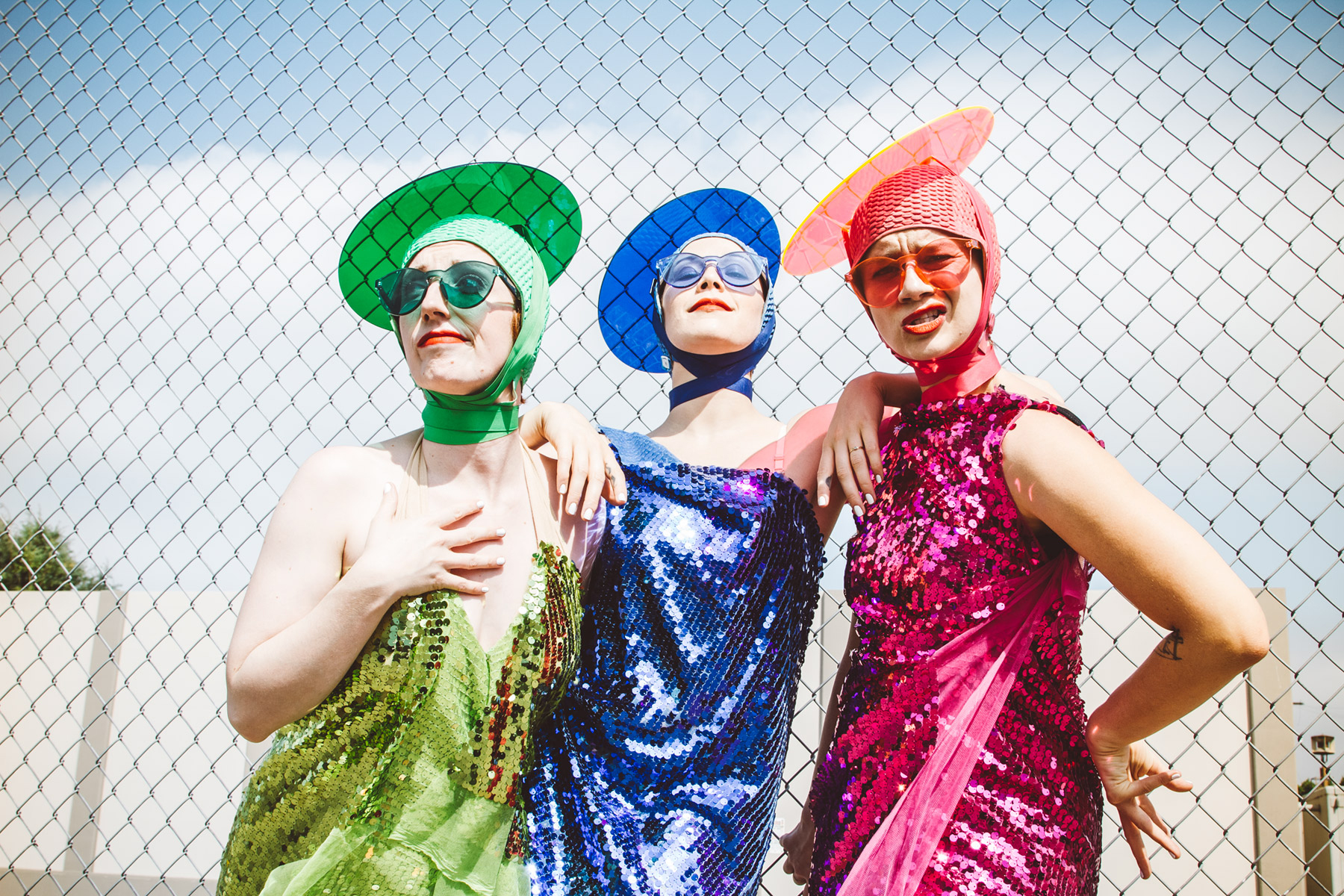 The three Seaweed Sisters strike quirky poses outdoors, backed by a blue sky. They wear sequined dresses, sunglasses, and disc-like hats; Meyers' ensemble is green, Wilson's is blue, and Lawson's is pink.
